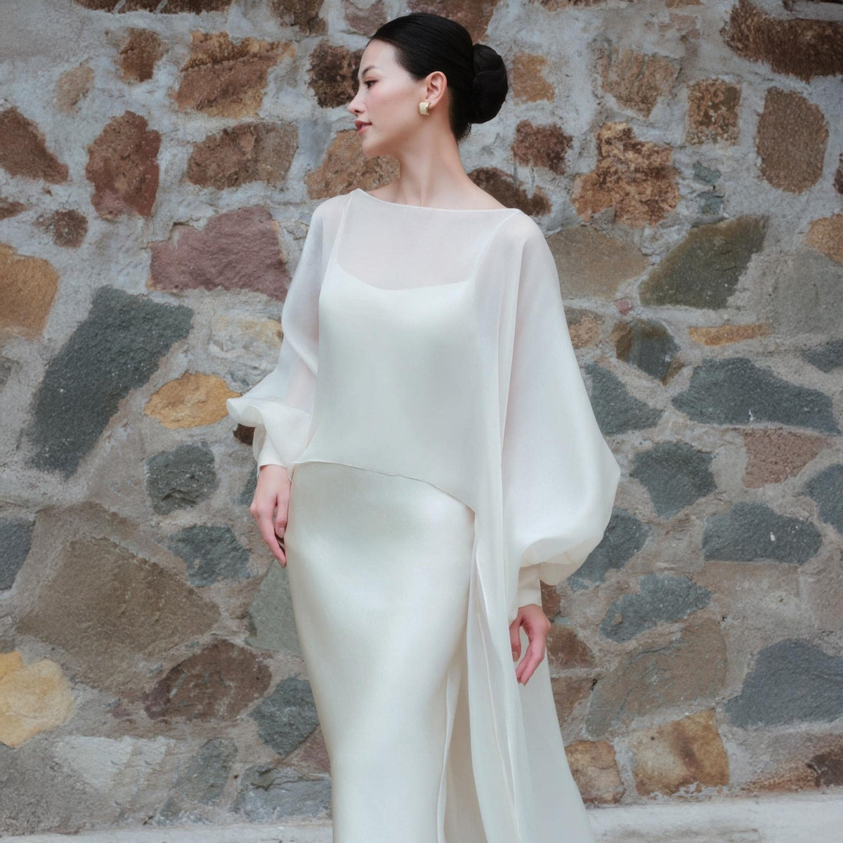 May include: A woman wearing a white wedding dress with a sheer, long-sleeved overlay. The dress has a simple, elegant design with a fitted bodice and a flowing skirt. The overlay features voluminous sleeves and a draped effect, creating a sophisticated look.
