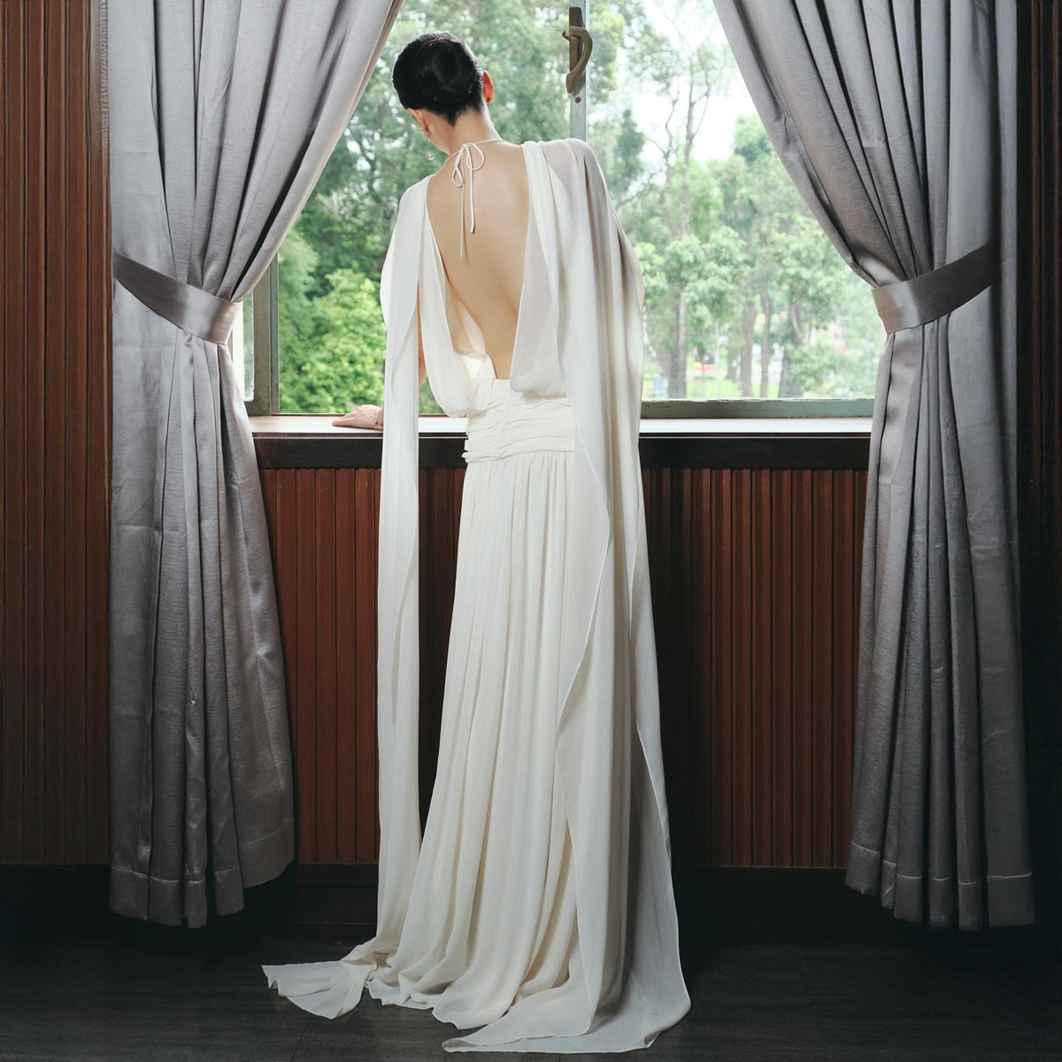 May include: A long, flowing ivory dress with a low back and draped sleeves. The dress features a gathered waist and a train that trails on the floor. The model is standing by a window with gray curtains, looking out at a green, tree-filled landscape.