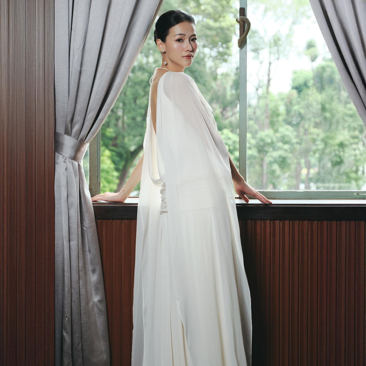 May include: A white, flowing gown with a cape-like design. The dress features a low back and a split in the front. The model is standing near a window with gray curtains and a wooden frame.