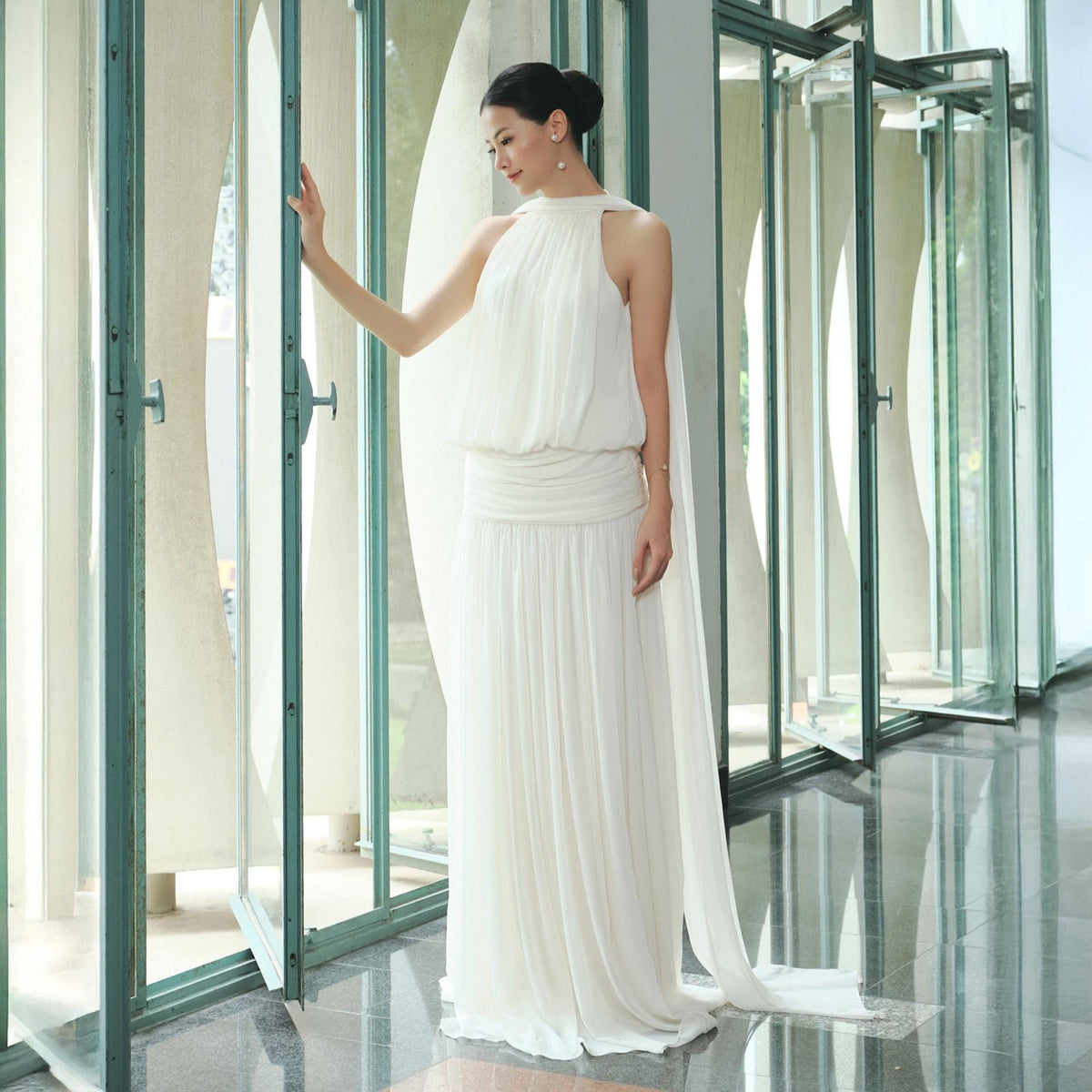 May include: A white halter-neck gown with a flowing skirt and a cape-like detail. The dress features a high neckline and a cinched waist. The model is standing near a large window with a modern architectural design.