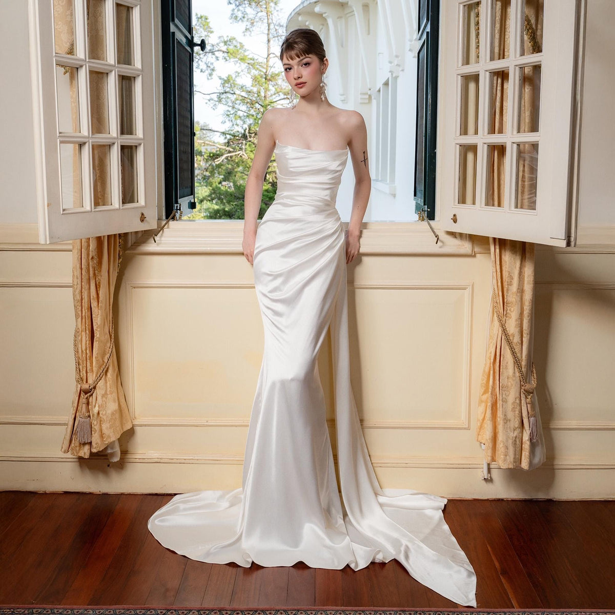 May include: A strapless, ivory-colored bridal gown with a mermaid silhouette. The dress features a draped bodice and a flowing train. The model is standing in front of a window with open shutters.