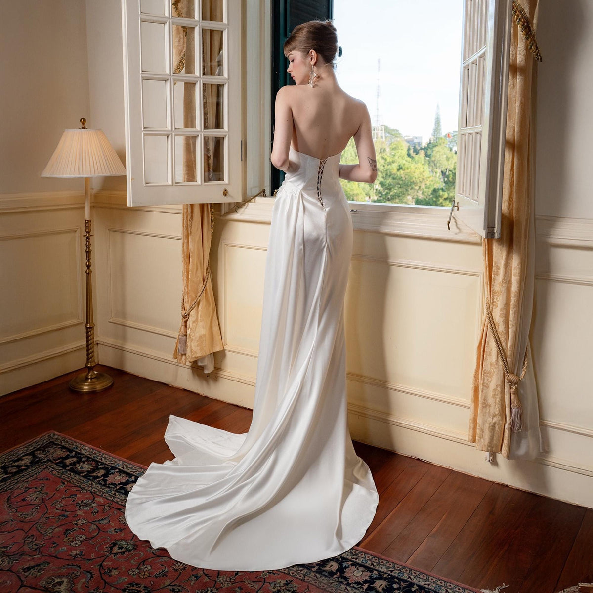 May include: A strapless, ivory-colored wedding gown with a flowing train. The dress features a fitted bodice with a corset back and a simple, elegant design. The bride is standing near a window with a view of trees and buildings.
