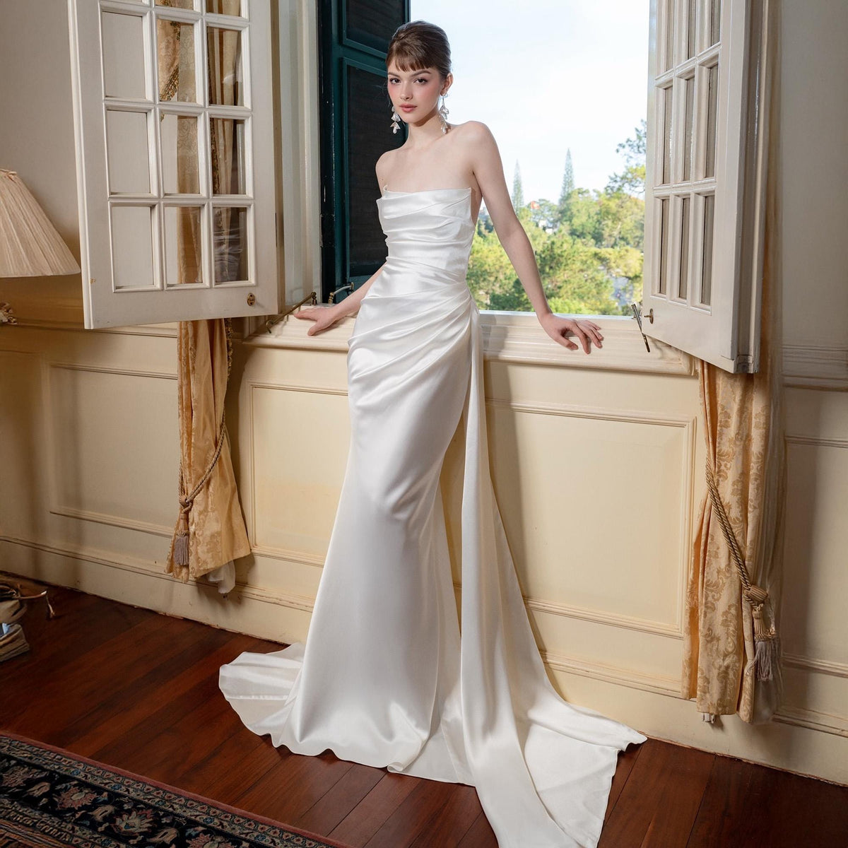 May include: A strapless white satin wedding gown with a draped bodice and a flowing train. The dress is floor-length and has a fitted silhouette. The model is standing near a window with a view of trees.