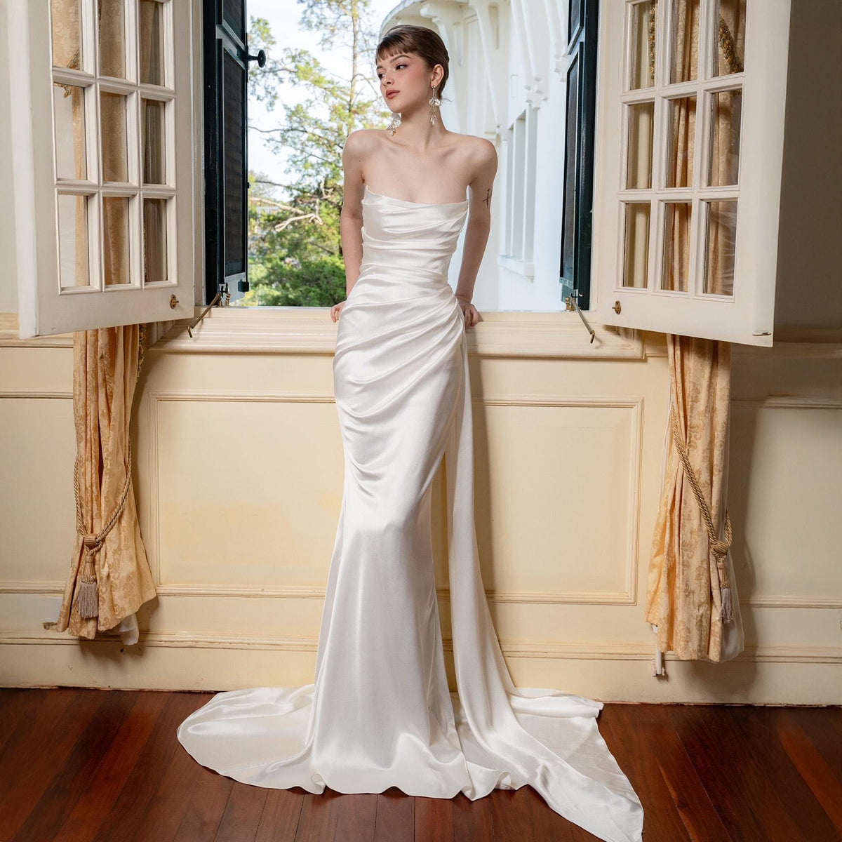 May include: A strapless, ivory-colored satin gown with a flowing train. The dress features a fitted bodice and a draped skirt, creating an elegant silhouette. The gown is displayed near an open window with natural light.