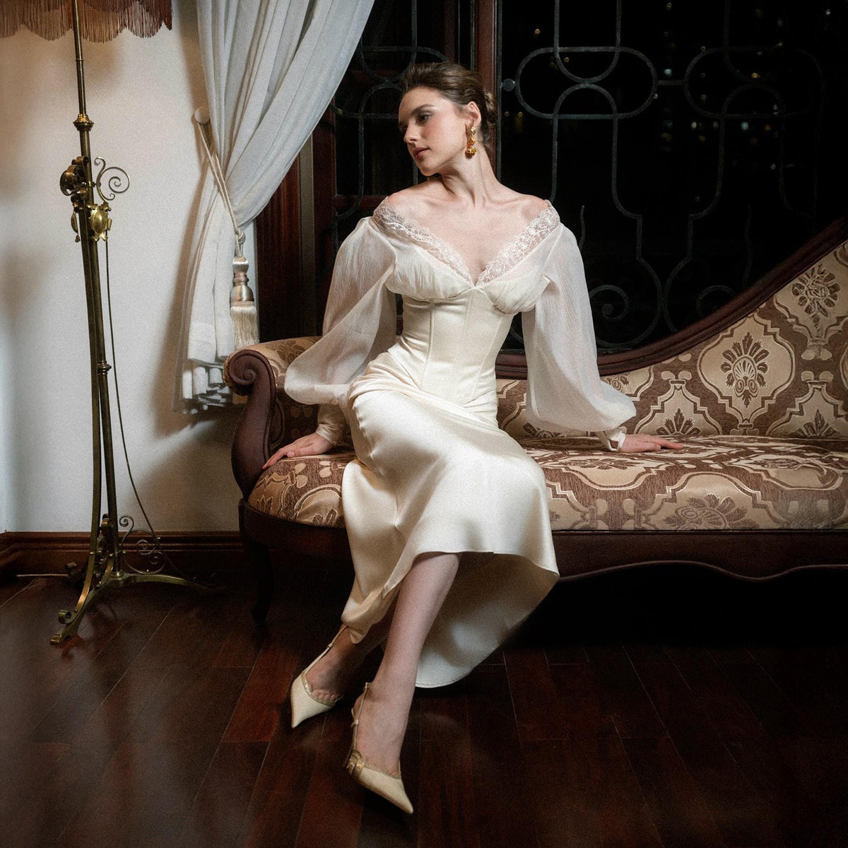May include: Ivory-colored satin dress with sheer, long sleeves and a lace-trimmed neckline. The dress features a fitted bodice and a flowing skirt. The model is wearing matching ivory-colored heels. The setting is a room with a vintage sofa and a floor lamp.