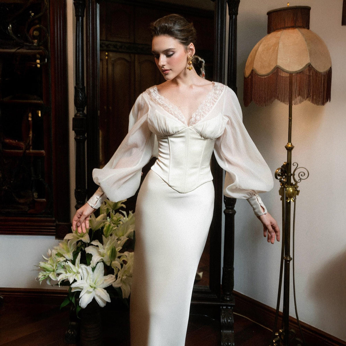 May include: Ivory-colored wedding dress with a fitted bodice and a long, flowing skirt. The dress features sheer, long sleeves and delicate lace detailing around the neckline. A bouquet of white lilies sits nearby.