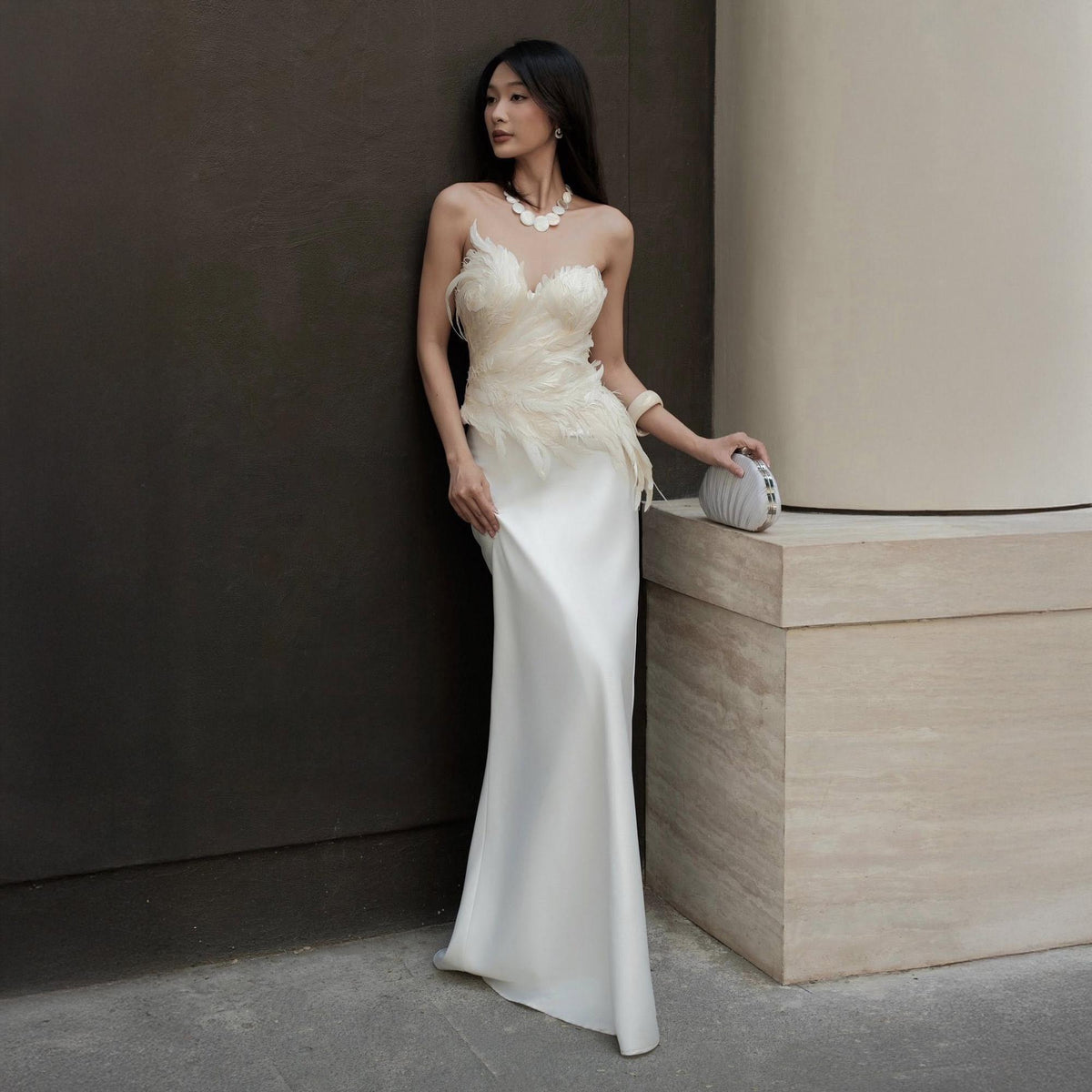 May include: A white, floor-length gown with a feathered bodice and a sleek, satin skirt. The dress features a strapless design and is paired with a small, silver clutch. The model is wearing a delicate necklace and bracelet.