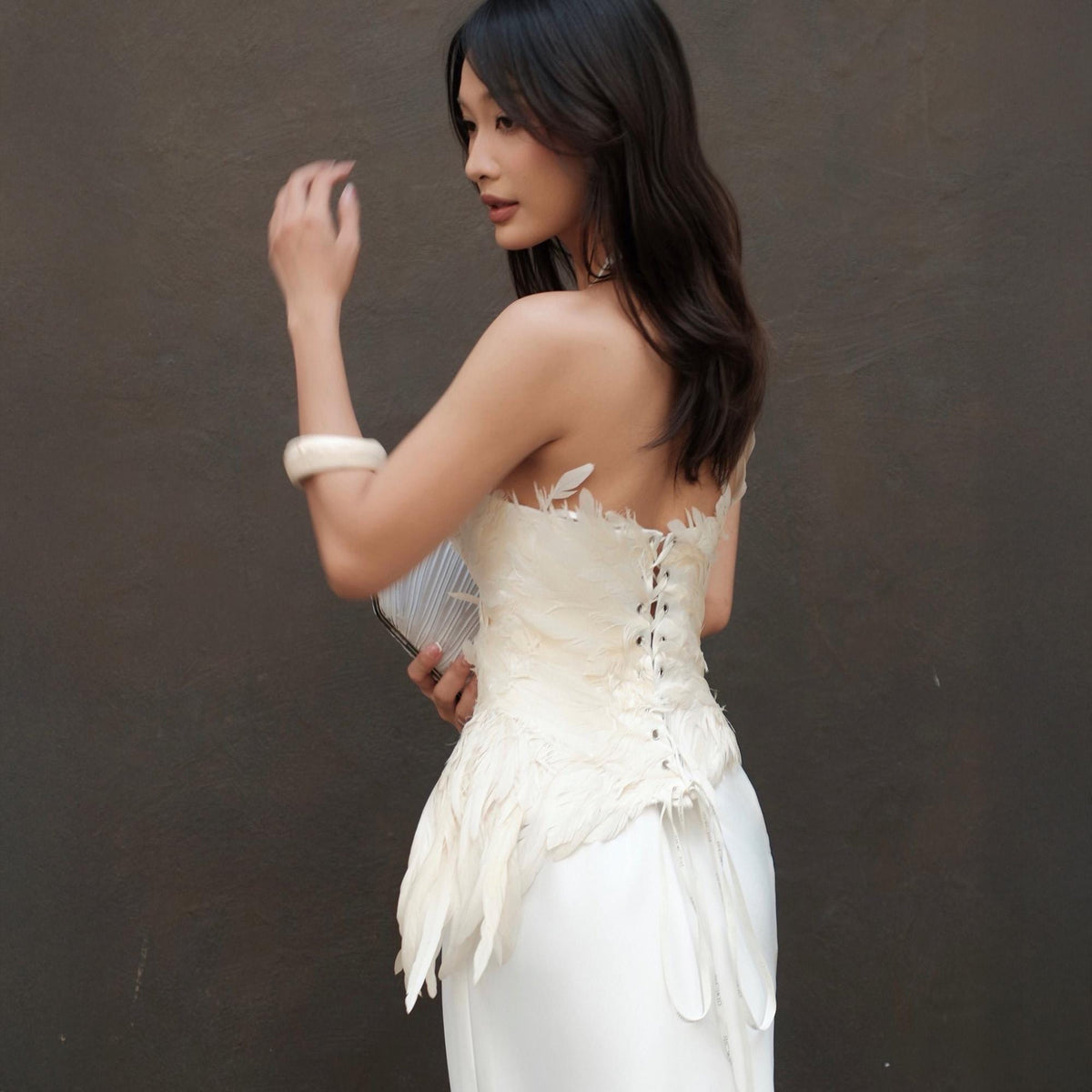 May include: A strapless white dress with a corset-style bodice adorned with feathers. The dress features a long, flowing skirt. A white bracelet and a silver clutch complete the look. The background is a solid, dark gray.
