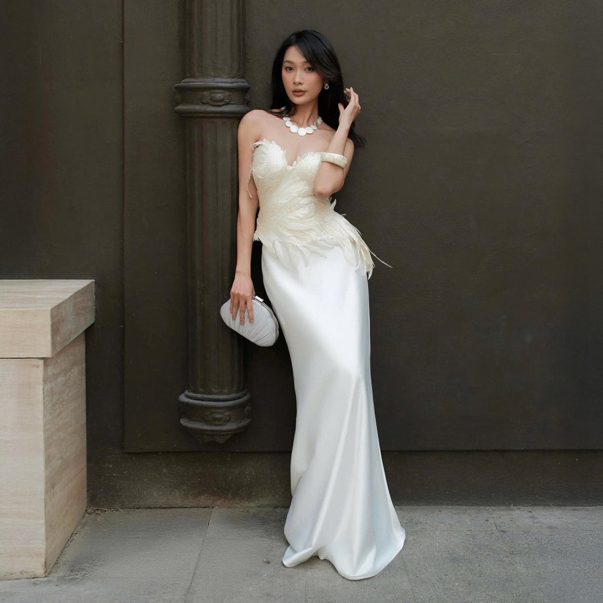 May include: A strapless, floor-length ivory gown with a feathered bodice and a sleek, flowing skirt. The dress is paired with a white clutch and a pearl necklace. The model is leaning against a dark wall.