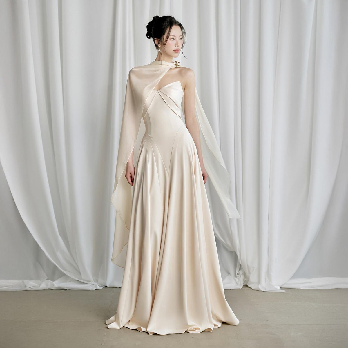 May include: A cream-colored formal gown with a flowing skirt and a draped, asymmetrical neckline. The dress features a cape-like overlay and a subtle, elegant design, suitable for special occasions.