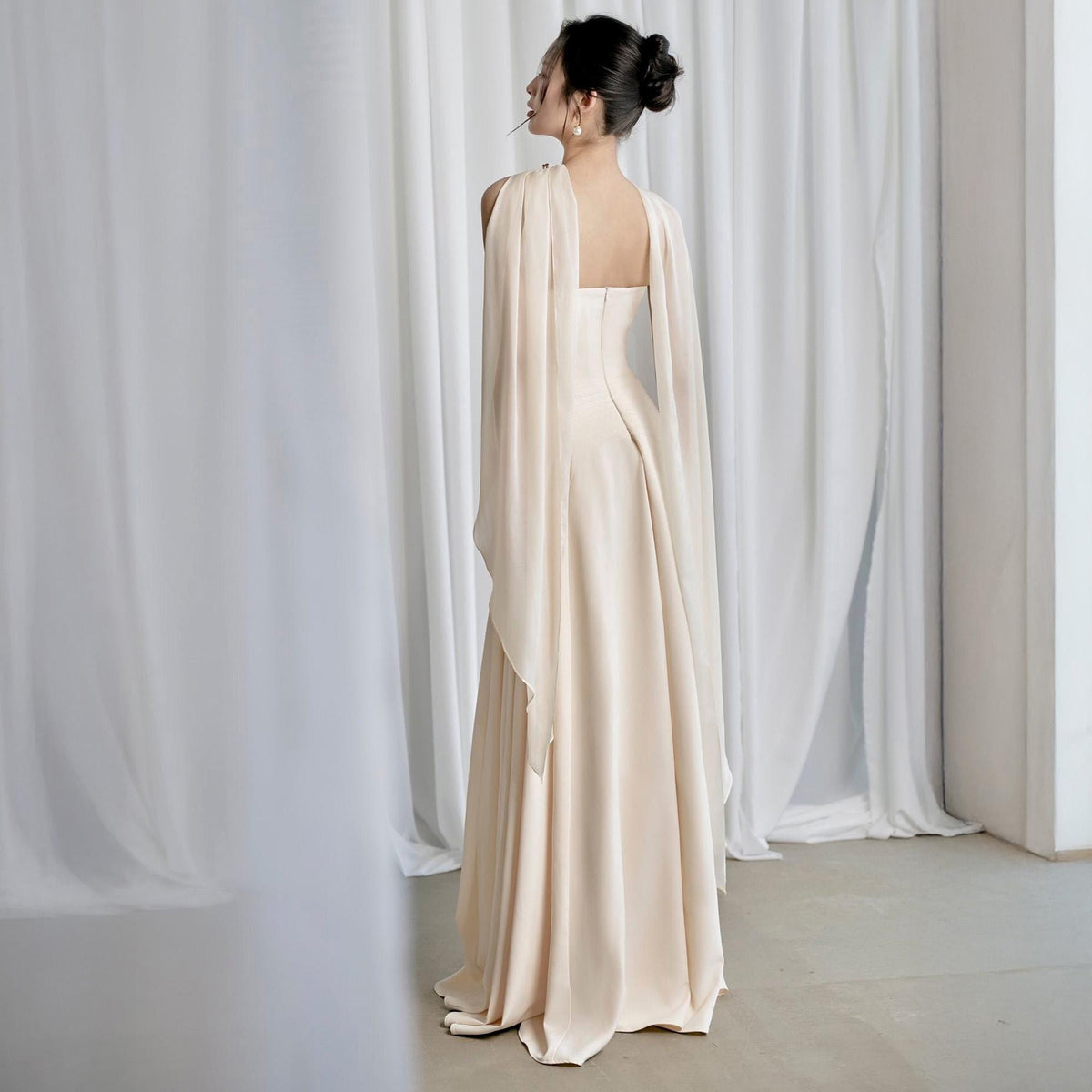 May include: A floor-length, cream-colored gown with a square neckline and flowing cape sleeves. The dress features a pleated skirt and a fitted bodice. The model is seen from the back, standing in front of white curtains.