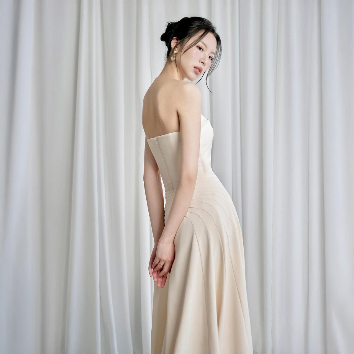 May include: A strapless, floor-length gown in a soft, creamy beige. The dress features a fitted bodice and a flowing skirt with subtle pleating details. The fabric appears smooth and elegant, suitable for formal events.
