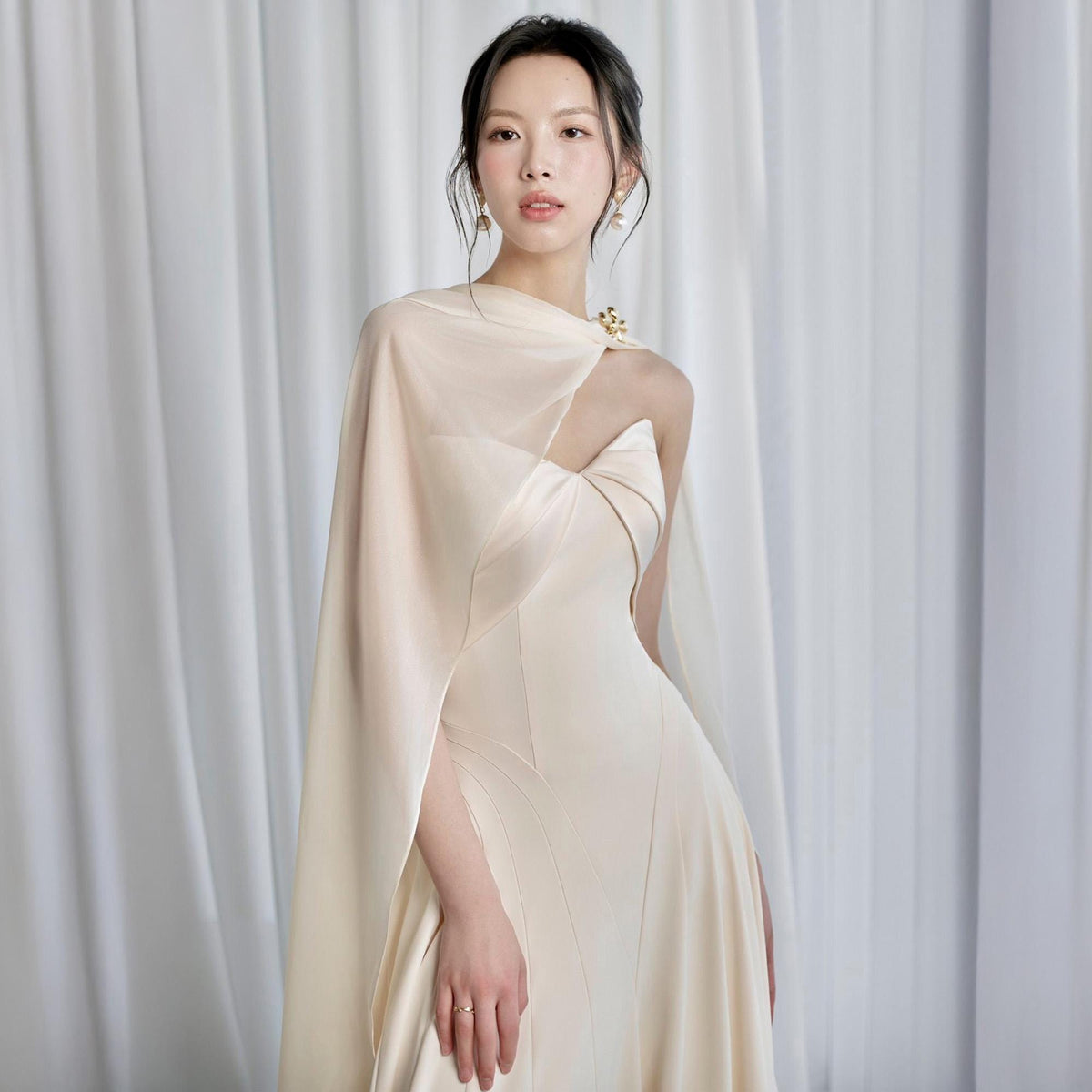 May include: A cream-colored formal gown with a cape. The dress features a fitted bodice and a flowing skirt. A gold-toned brooch secures the cape at the shoulder. The gown is made of a smooth, possibly satin, fabric.