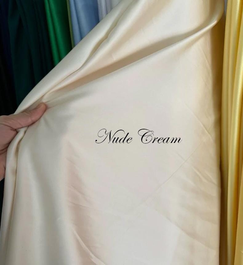 May include: A close-up of a cream-colored fabric with the text "Nude Cream" in elegant script. The fabric is draped, showing its smooth texture and subtle sheen. Other fabrics in green and yellow are visible in the background.
