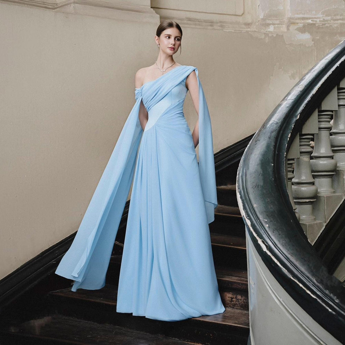 May include: A light blue, one-shoulder formal gown with flowing cape sleeves. The dress features a fitted bodice and a floor-length skirt. The model is standing on a wooden staircase with a dark railing.