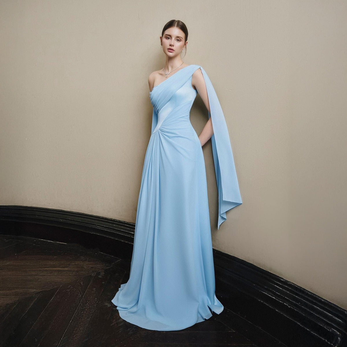 May include: A light blue, one-shoulder formal gown with a flowing cape detail. The dress features a draped bodice and a floor-length skirt. The model is standing on a wooden floor with a neutral-colored wall in the background.