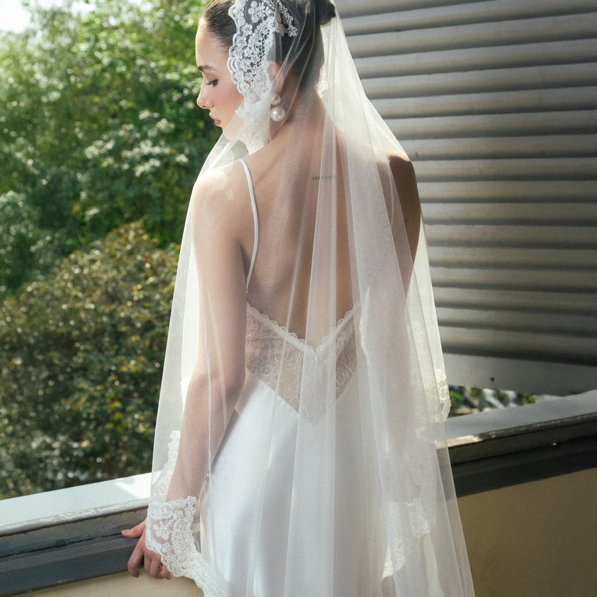May include: A bride wearing a white satin wedding dress with a low back and delicate lace detailing. She is wearing a long, sheer veil and a lace headpiece. The bride is standing on a balcony with greenery in the background.