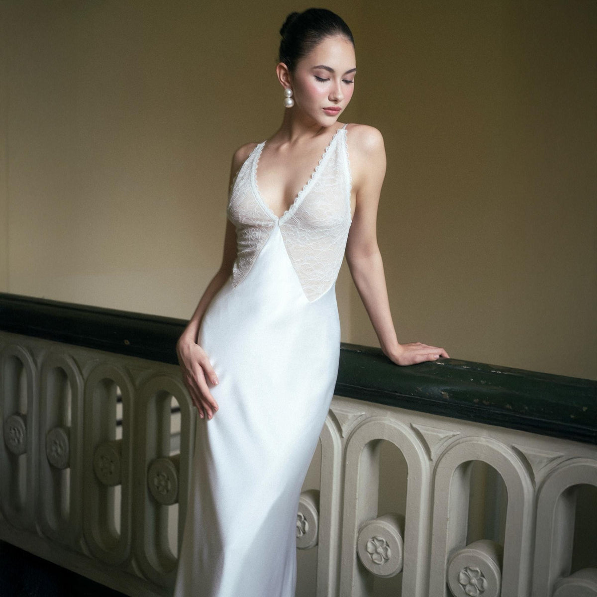 May include: A white satin slip dress with a lace bodice. The dress features a V-neckline and spaghetti straps. The model is wearing pearl earrings. The background is a neutral color.