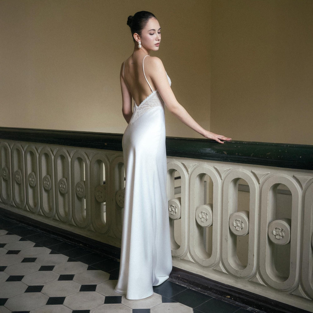 May include: A white satin slip dress with a low back and spaghetti straps. The dress features lace detailing on the bodice and a flowing skirt. The model is standing on a black and white tiled floor, leaning on a decorative railing.
