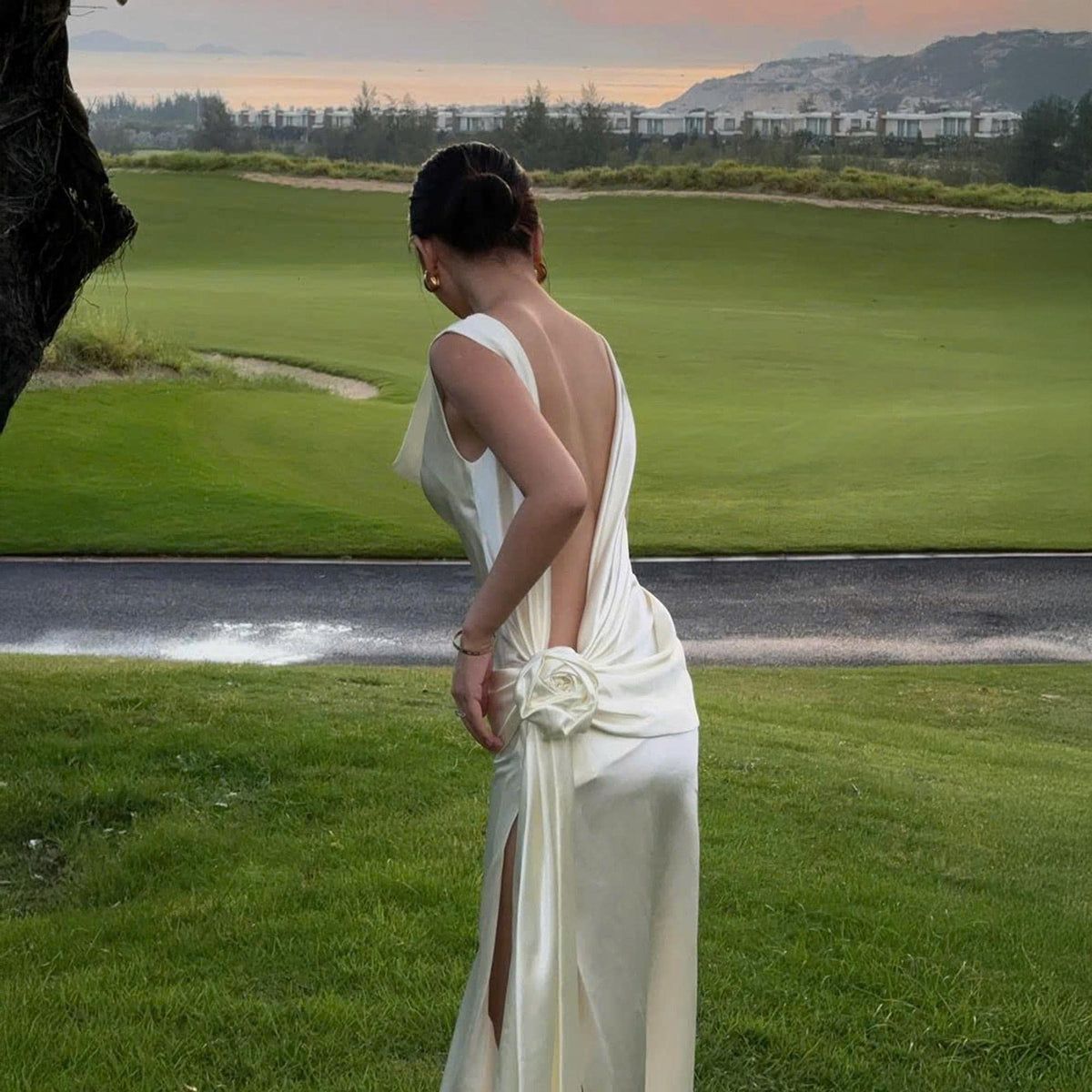 May include: A white satin gown with a low back and a high slit. The dress features a rose detail at the waist and a flowing skirt. The background shows a green golf course and a view of the ocean.
