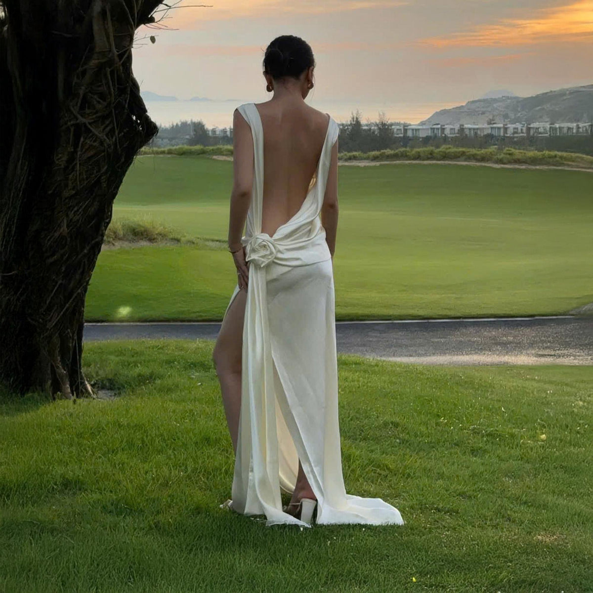 May include: A cream-colored, backless formal gown with a high slit and a floral accent at the waist. The dress has a flowing train and is worn outdoors on a green lawn with a golf course in the background.