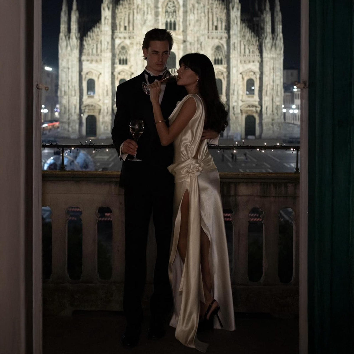 May include: A couple in formal attire, a tuxedo and a satin gown, share a moment on a balcony overlooking a cathedral at night. The woman's dress has a high slit and a floral detail at the waist. They are holding wine glasses.