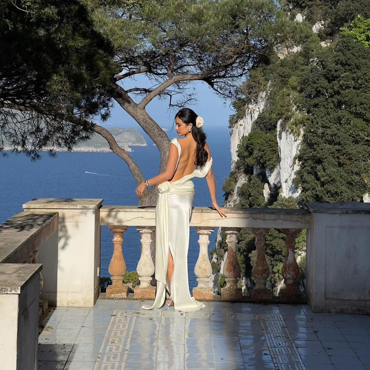 May include: A cream-colored, backless formal gown with a high slit. The dress features a draped design and a floral accent at the waist. The person is standing on a balcony overlooking the ocean, with a tree and mountains in the background.