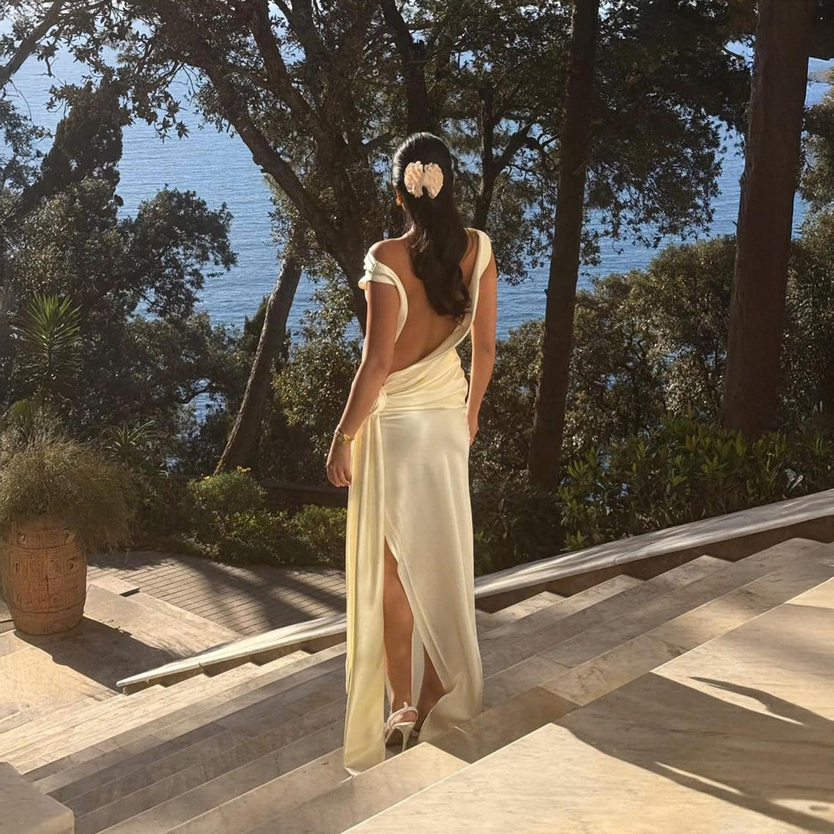 May include: A woman in a cream-colored, backless dress with a high slit, walking down stone steps. Her dark hair is styled with a decorative hairpiece. The background features trees, foliage, and a view of the ocean.