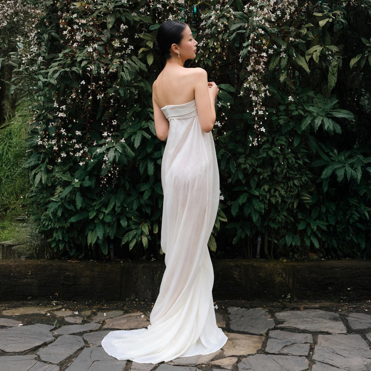 May include: A strapless, floor-length white gown with a flowing train. The dress is made of a sheer, lightweight fabric. The background features lush green foliage and white flowers.