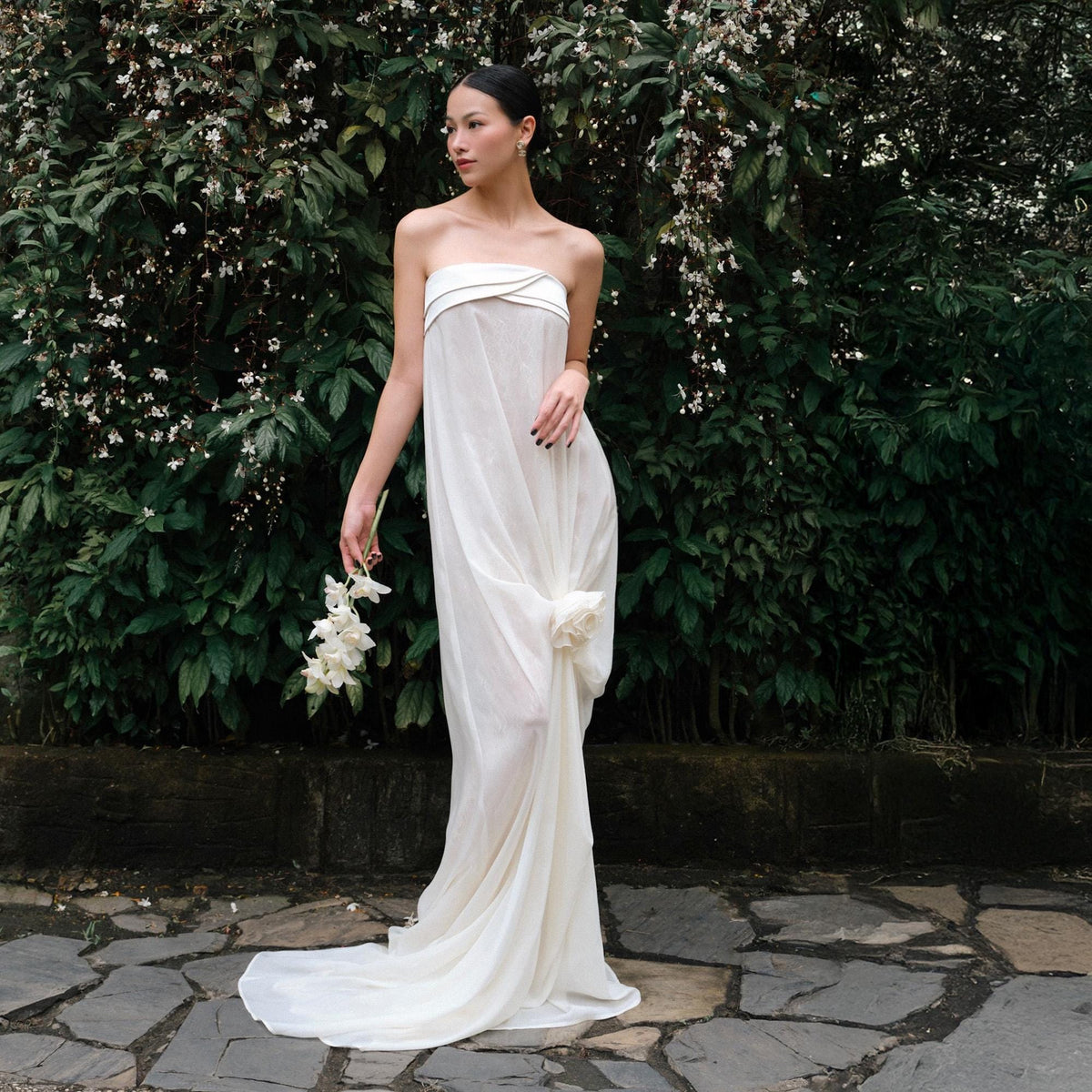 May include: A strapless white gown with a flowing skirt and a gathered detail at the waist. The dress is made of a lightweight fabric and has a subtle train. The model is holding a bouquet of white flowers.