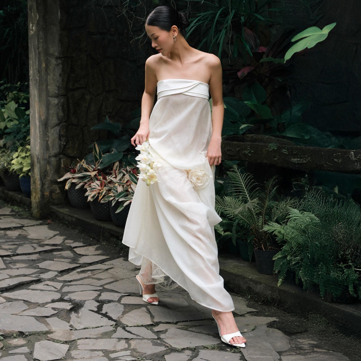 May include: A strapless ivory gown with a flowing skirt and floral accents. The dress features a draped bodice and is paired with white heels. The setting is an outdoor garden with lush greenery and a stone pathway.