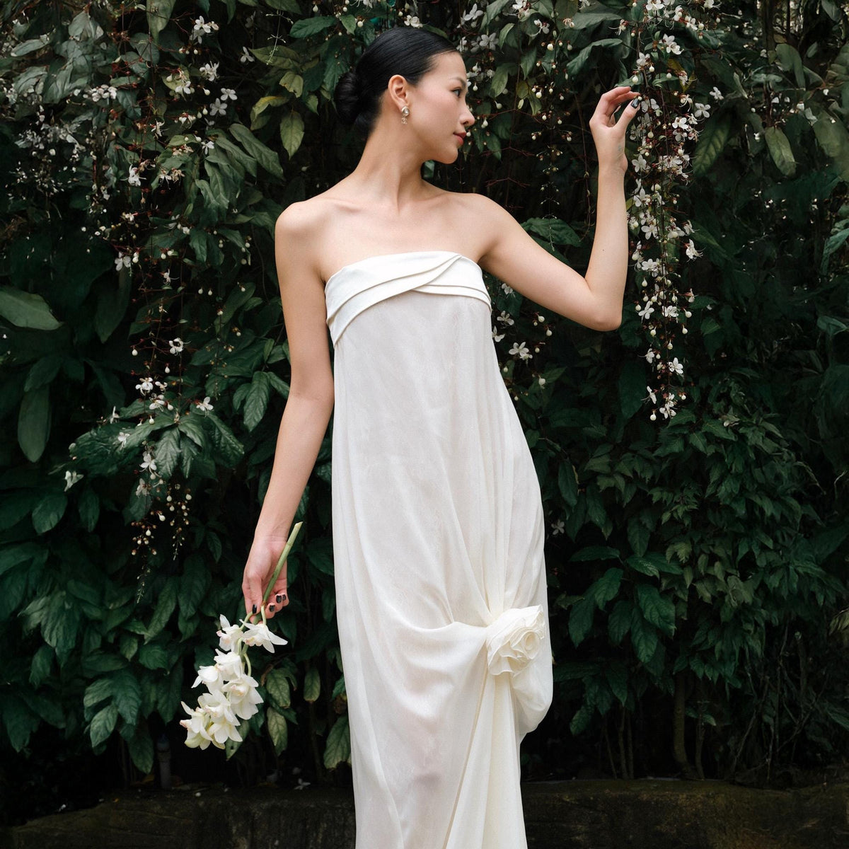 May include: A strapless white dress with a flowing silhouette. The dress features a decorative knot detail at the hip and a contrasting trim at the neckline. The person is holding white flowers, set against a backdrop of green foliage.