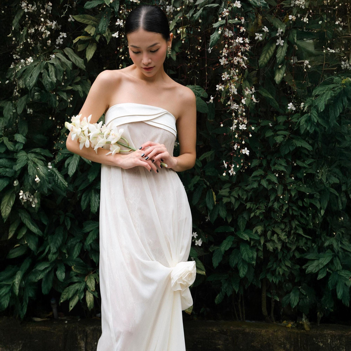 May include: A strapless white dress with a draped skirt and a bow detail. The dress is paired with a bouquet of white flowers. The background features lush green foliage and white flowers.