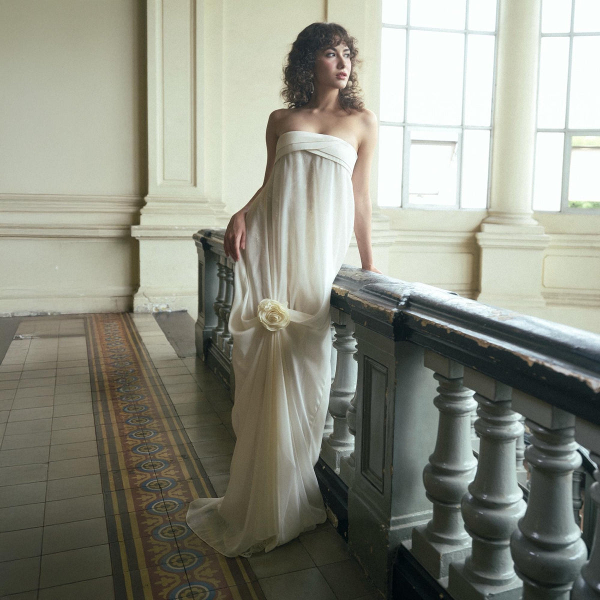 May include: A strapless, floor-length ivory gown with a draped design and a floral accent at the waist. The dress is worn by a person standing on a balcony with a decorative railing.