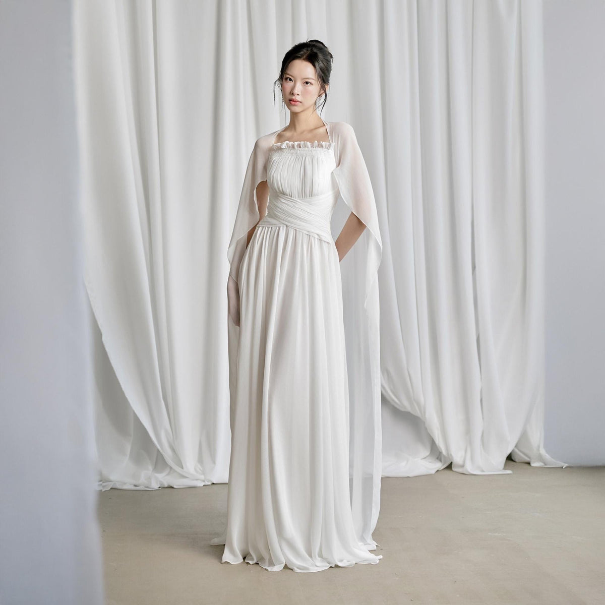 May include: A white, floor-length gown with a flowing skirt and a fitted bodice. The dress features a sheer, cape-like overlay that drapes over the shoulders. The model is standing in front of white curtains.