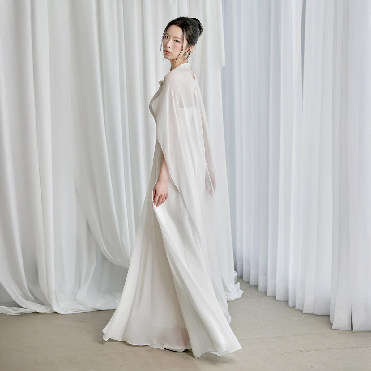 May include: A flowing, floor-length white gown with a sheer cape. The dress features a high neckline and a simple, elegant design. The model is standing in front of white curtains, creating a soft, ethereal look. The gown is suitable for formal events.