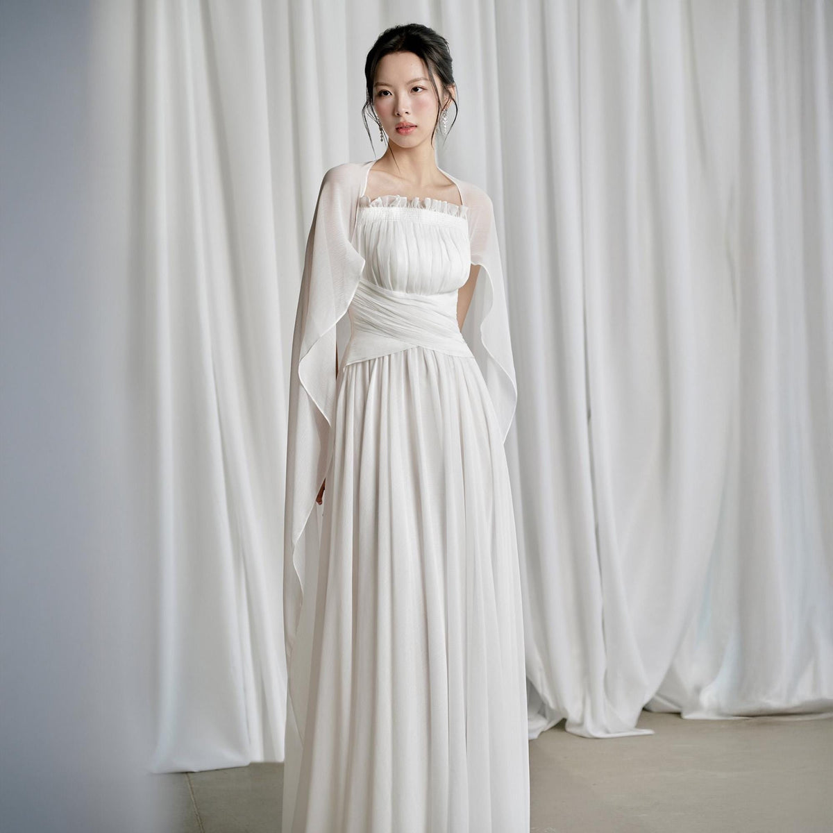 May include: A white formal gown with a square neckline and a flowing skirt. The dress is paired with a sheer, matching shawl. The fabric appears to be lightweight and drapes elegantly. The background is a white curtain.