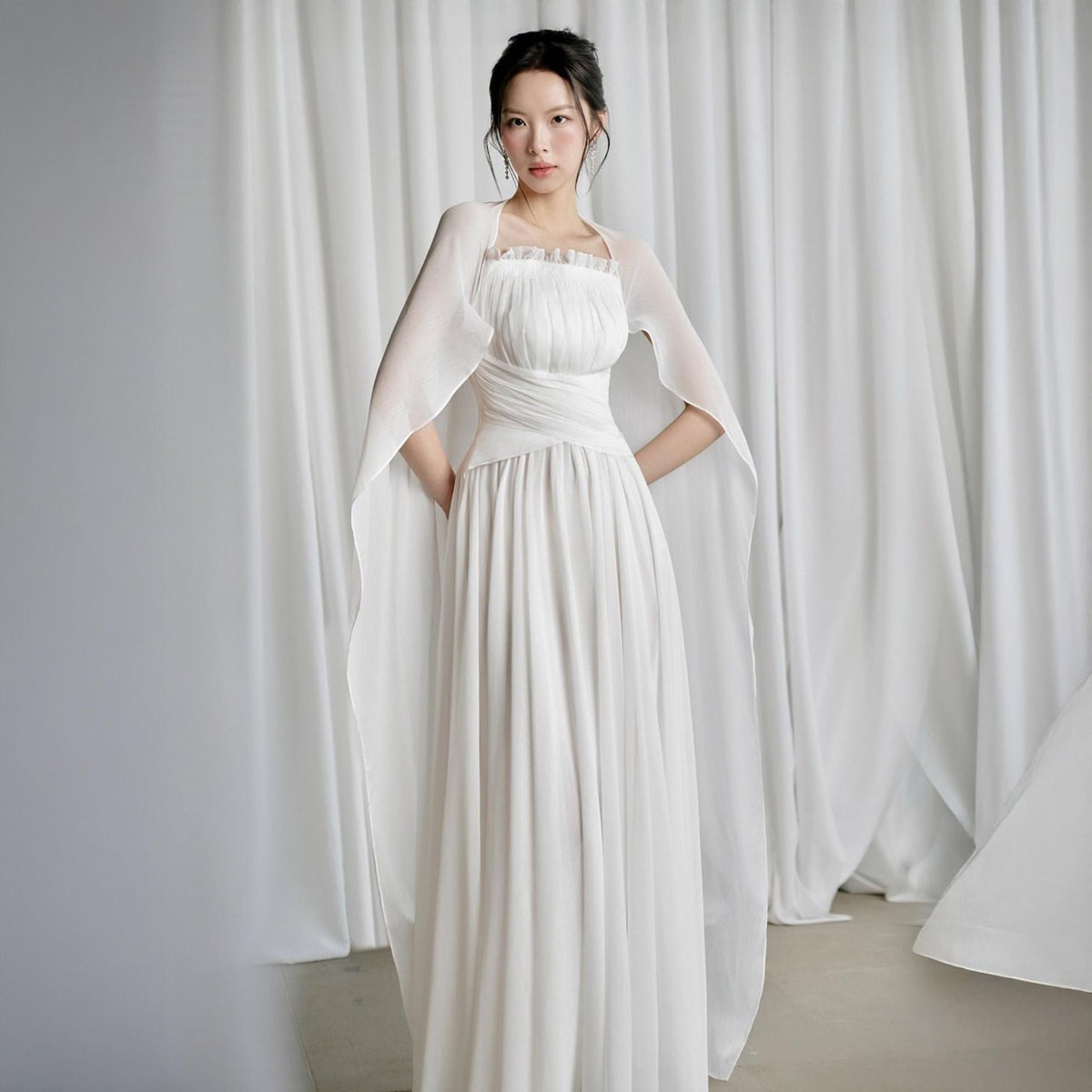 May include: A white, floor-length gown with a fitted bodice and flowing skirt. The dress features a draped waist and is paired with a sheer, cape-like overlay. The model is standing in front of white curtains.