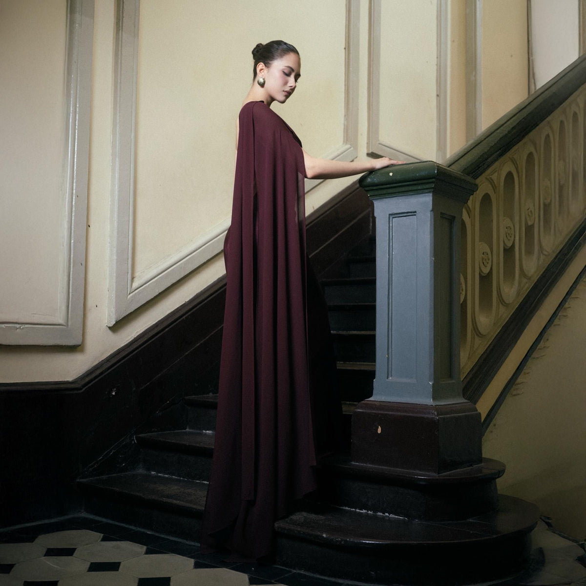 May include: A burgundy cape dress with a flowing design. The dress is worn on a staircase with a dark wood banister and black steps. The background features cream-colored walls and a patterned floor.
