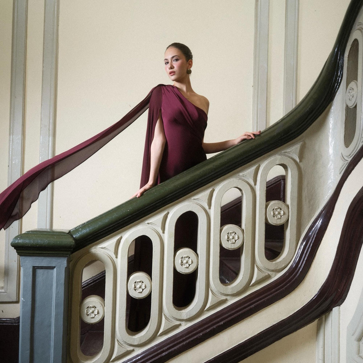 May include: A burgundy one-shoulder dress with a flowing cape detail. The dress is worn by a person standing on a staircase with a green and white banister. The staircase has decorative carved elements.