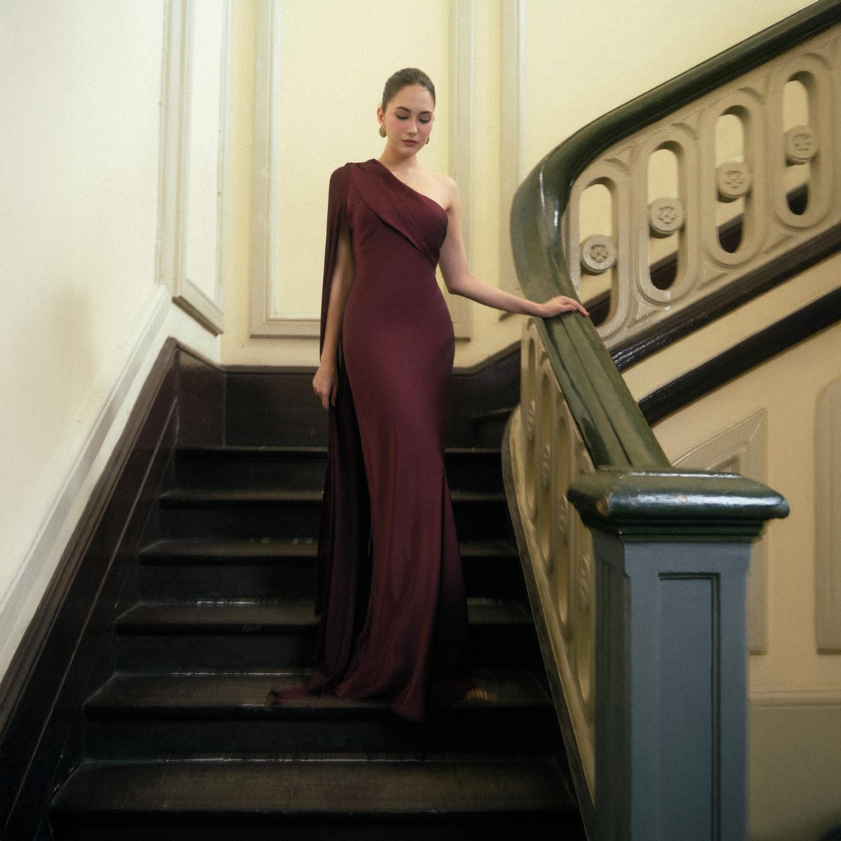 May include: A burgundy one-shoulder evening gown with a flowing cape detail. The dress is floor-length and form-fitting, with a draped neckline. The model is standing on a dark wooden staircase with a decorative banister.