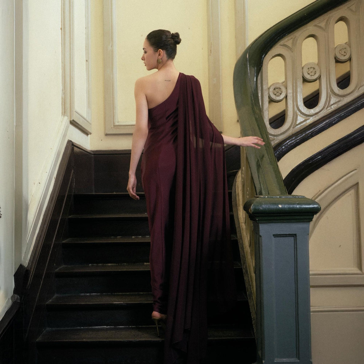May include: A burgundy one-shoulder draped dress. The dress is flowing and elegant, with a long train. The model is walking up a dark wooden staircase with a decorative banister. The background is a light-colored wall.