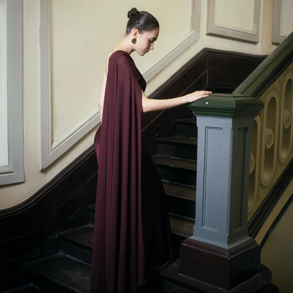 May include: A burgundy evening gown with a flowing cape detail. The dress is worn by a person descending a wooden staircase with a gray and green banister. The background is a light beige color.