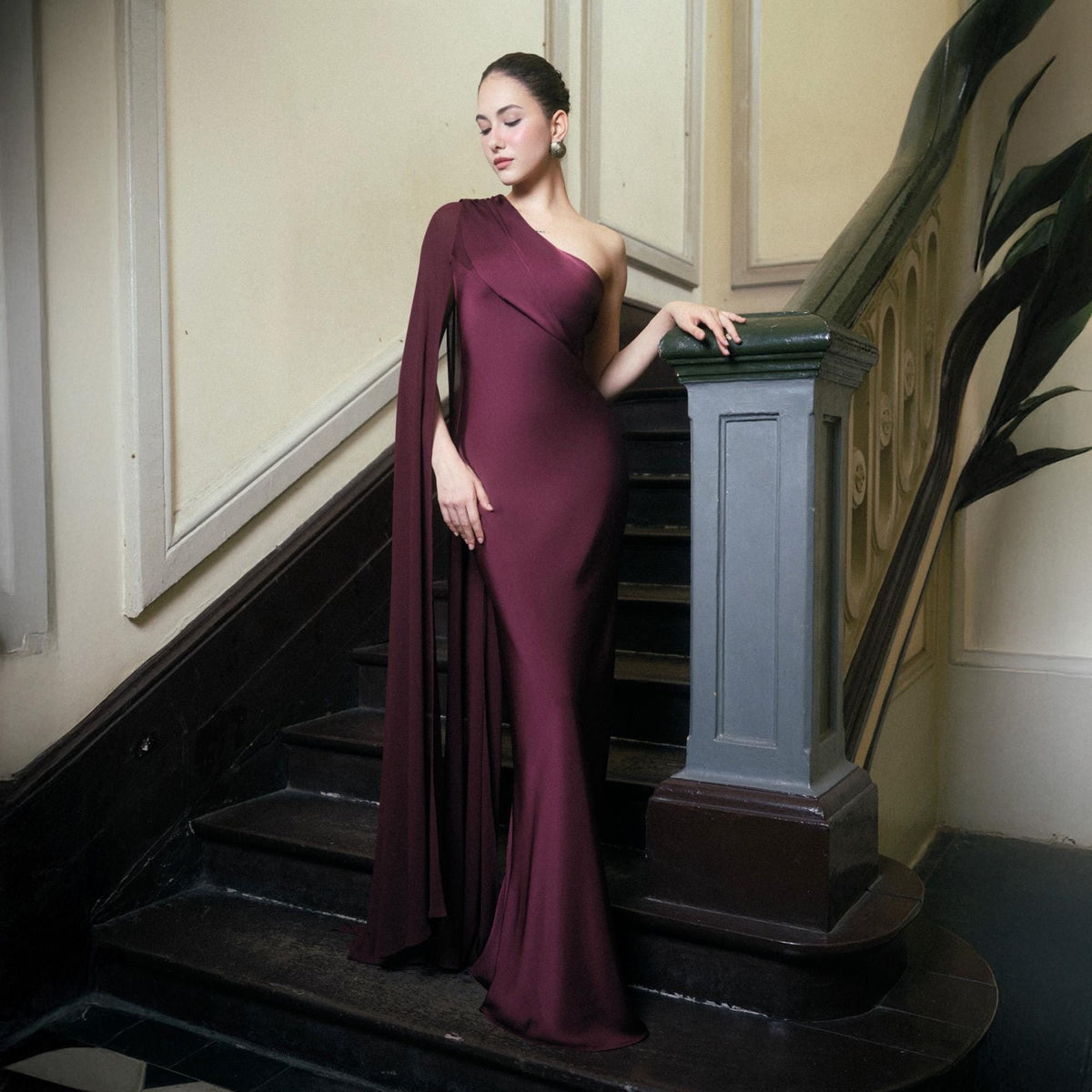 May include: A burgundy one-shoulder gown with a flowing cape. The dress is form-fitting, with a mermaid silhouette. The model is standing on a staircase with a dark wood banister. The dress is a deep, rich color.