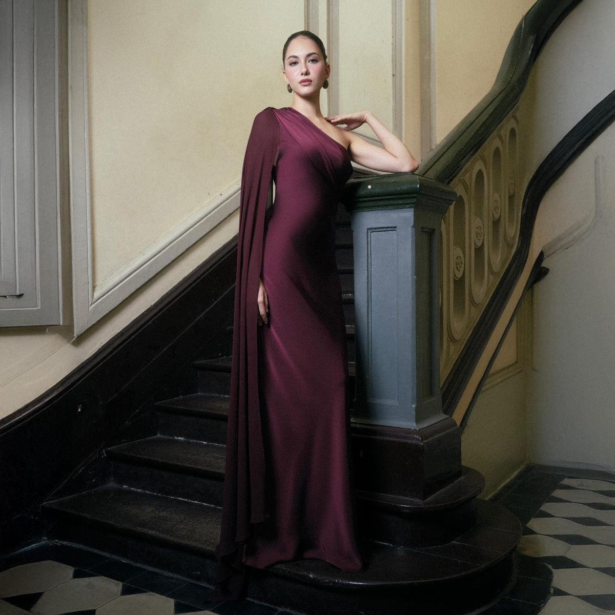 May include: A burgundy one-shoulder gown with a flowing cape sleeve. The dress is floor-length and made of a smooth, draping fabric. The model is standing on a staircase with a dark wood banister.