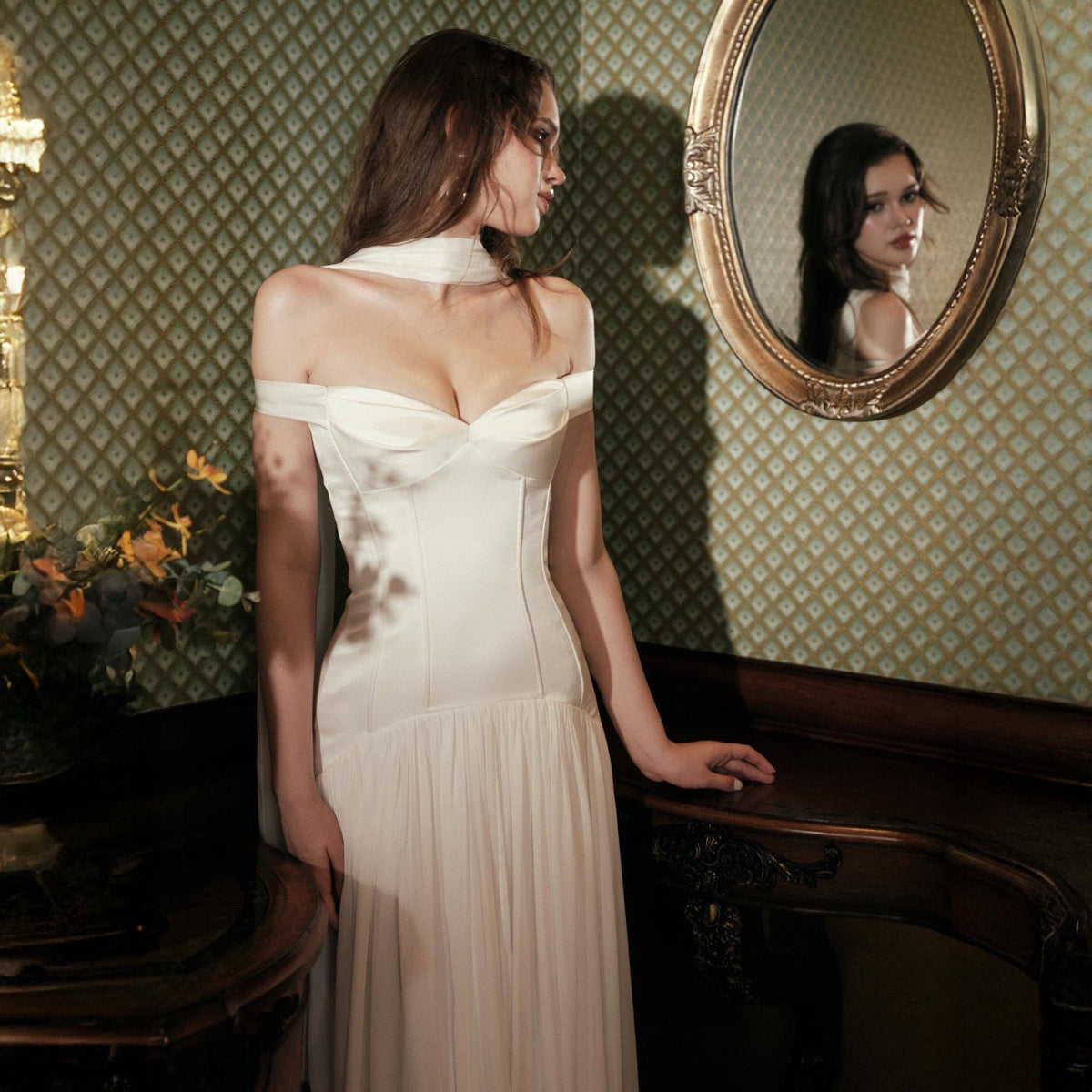 May include: An off-the-shoulder ivory gown featuring a sweetheart neckline, a structured bodice with vertical seams, and a flowing skirt. The garment includes a matching scarf draped around the neck, creating a cohesive look with a smooth satin finish.