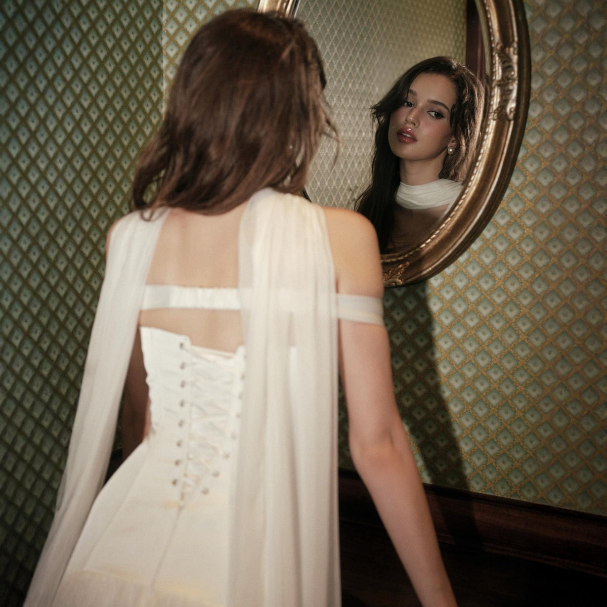 May include: A white strapless gown featuring a corset-style back with lace-up ribbon closure and long, sheer, flowing fabric panels draped over the shoulders. The garment is captured from behind, showing the bodice structure and fabric texture.