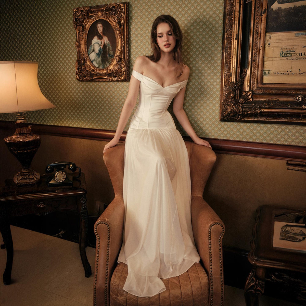 May include: An off-the-shoulder ivory gown featuring a structured corset bodice with a sweetheart neckline and a flowing, floor-length skirt made of sheer, lightweight fabric that drapes elegantly over the seat of a brown leather armchair.