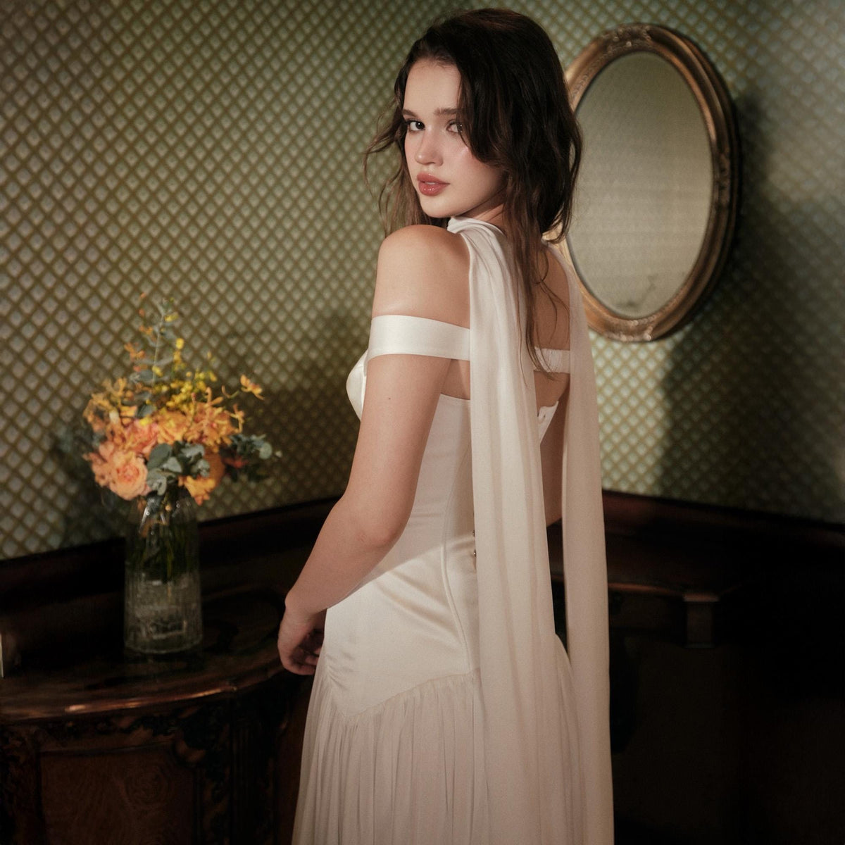 May include: A floor-length white gown featuring an off-the-shoulder neckline with wide, flowing fabric straps that drape down the back. The bodice is fitted with a smooth finish, transitioning into a pleated skirt, creating a structured silhouette.