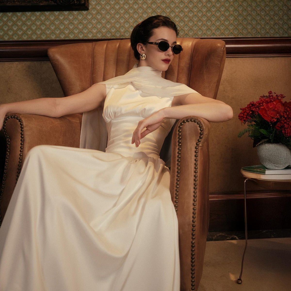 May include: A woman wearing a cream-colored satin gown and sunglasses sits in a brown leather armchair. The dress has a fitted bodice and a flowing skirt. Red flowers in a small vase sit on a side table.